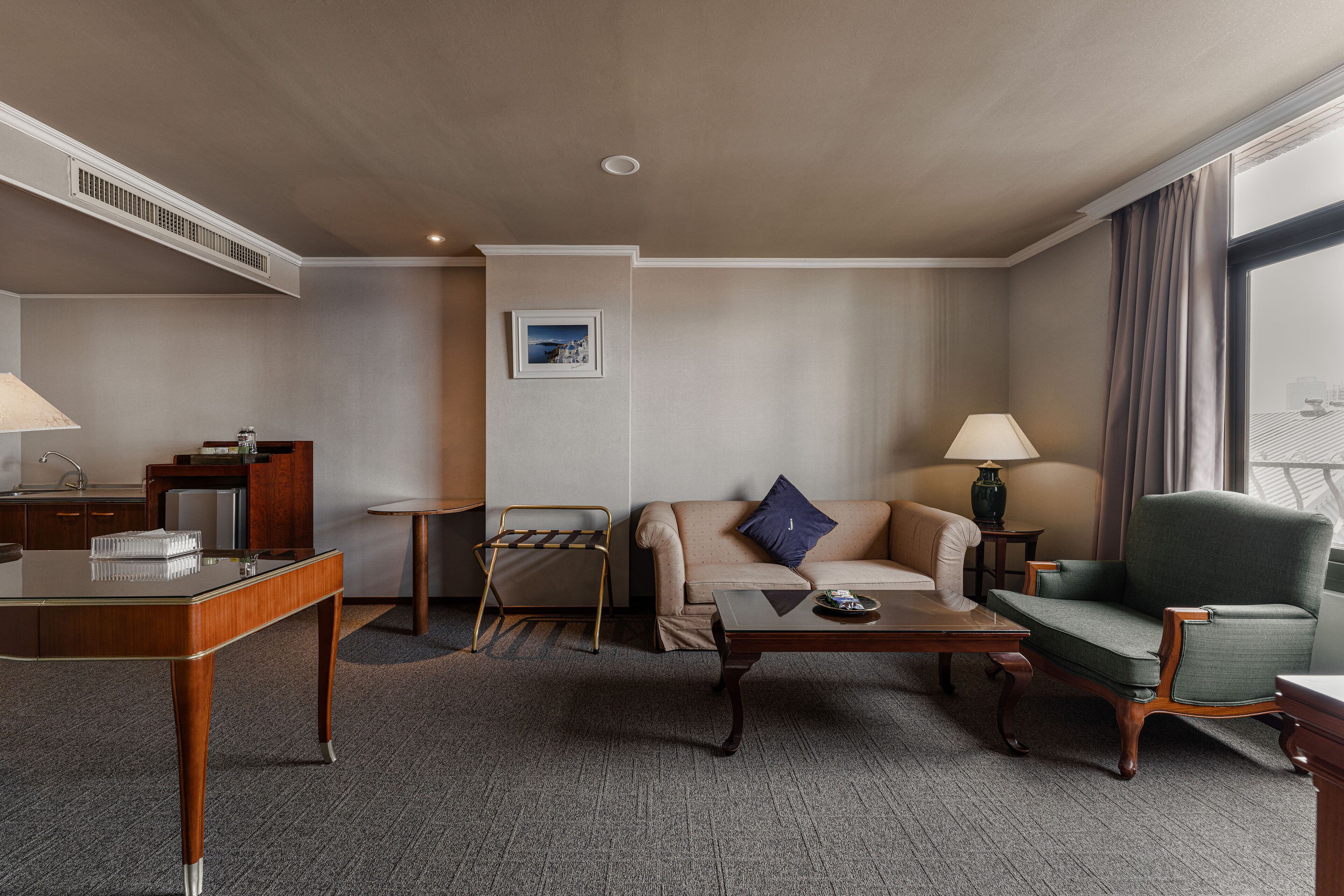 A hotel room with a sofa, armchair, coffee table, and a desk with a lamp.
