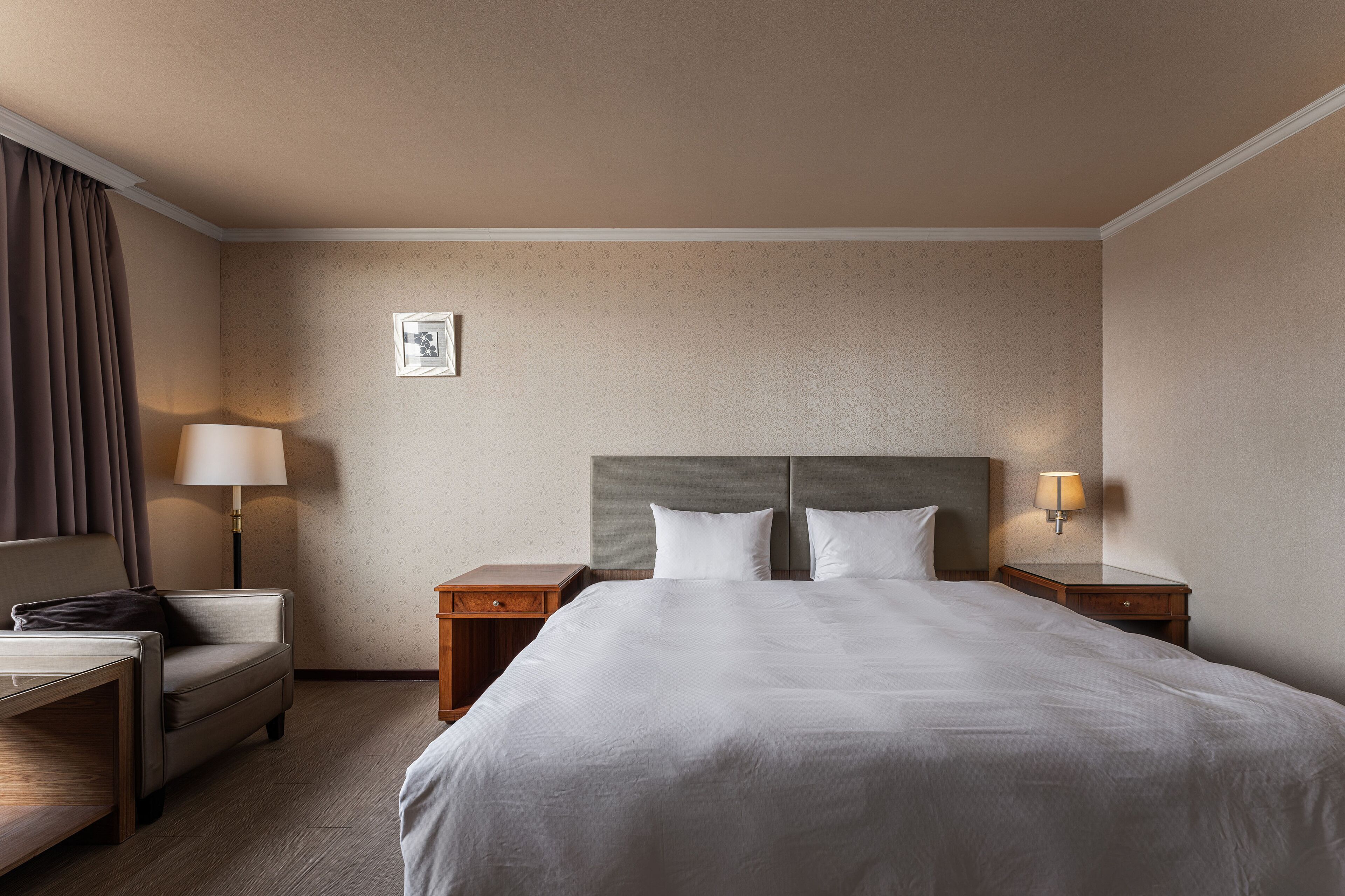 Ein Hotelzimmer mit einem großen Bett, zwei Nachttischen mit Lampen, einem kleinen Schreibtisch mit Stuhl und einem großen Fenster mit Vorhängen.