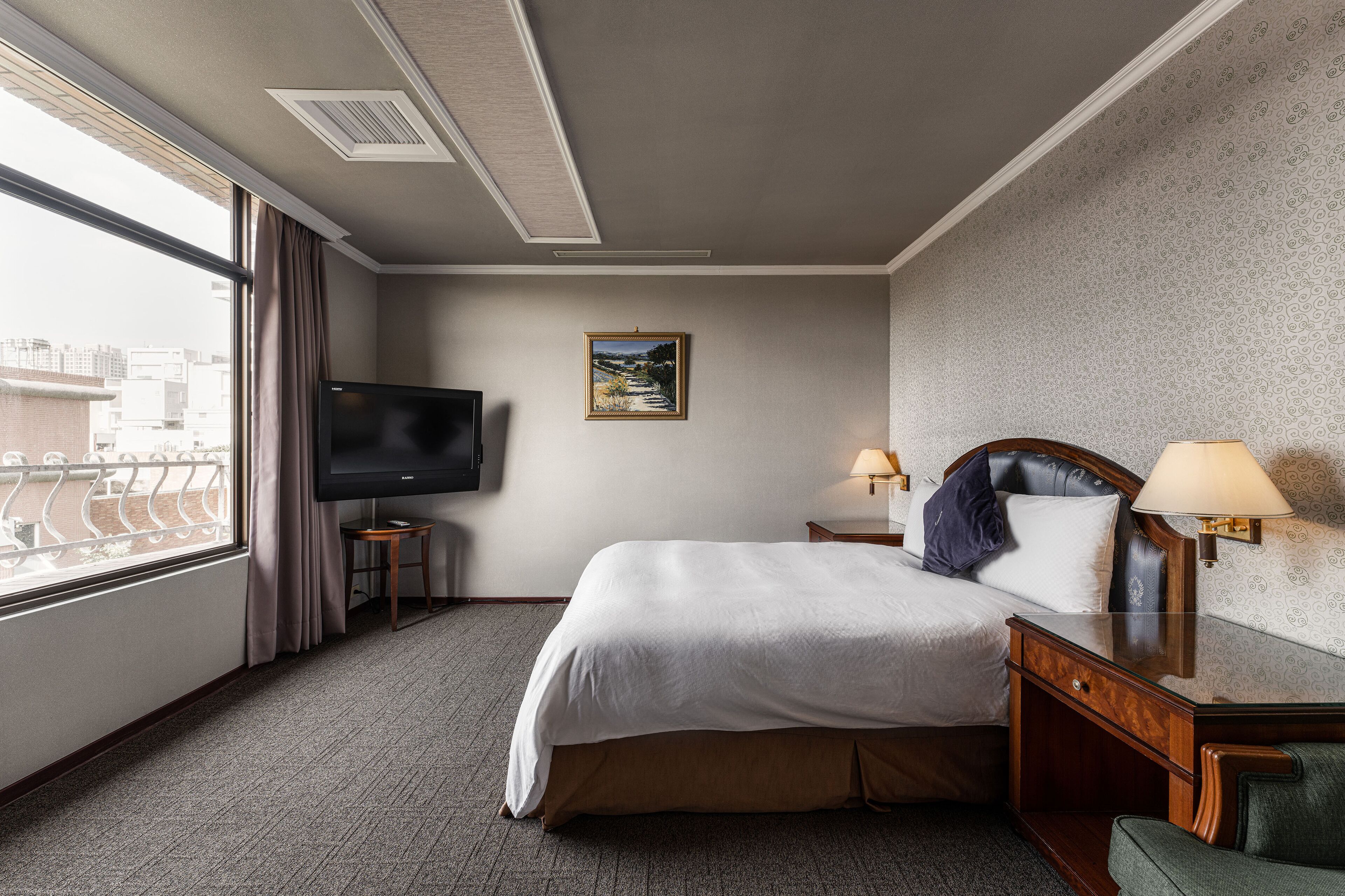 Una habitación de hotel con cama, televisión, ventana con cortinas, una mesita pequeña y una lámpara.