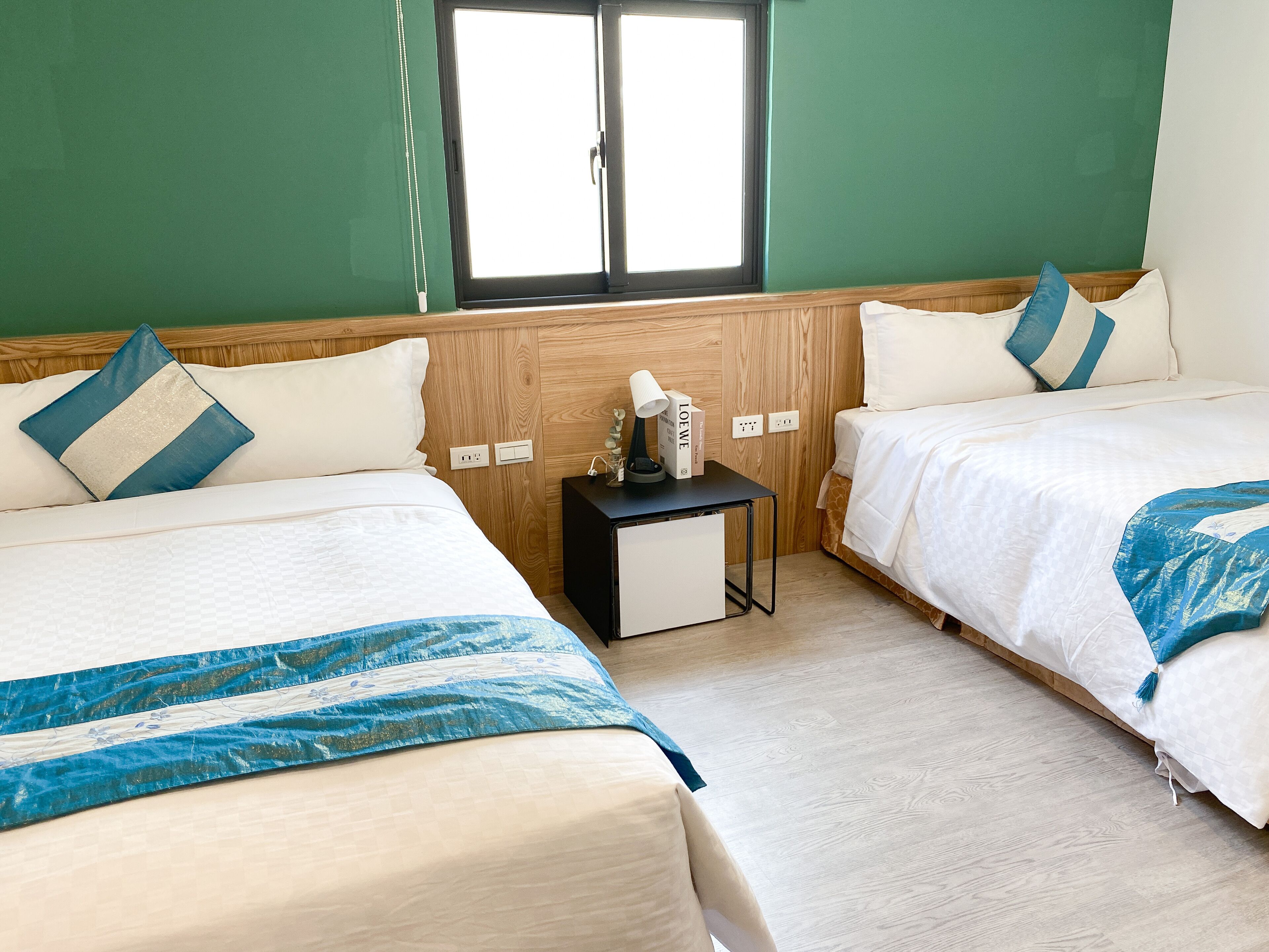 A hotel room with two beds, a small table with a lamp, and a window.