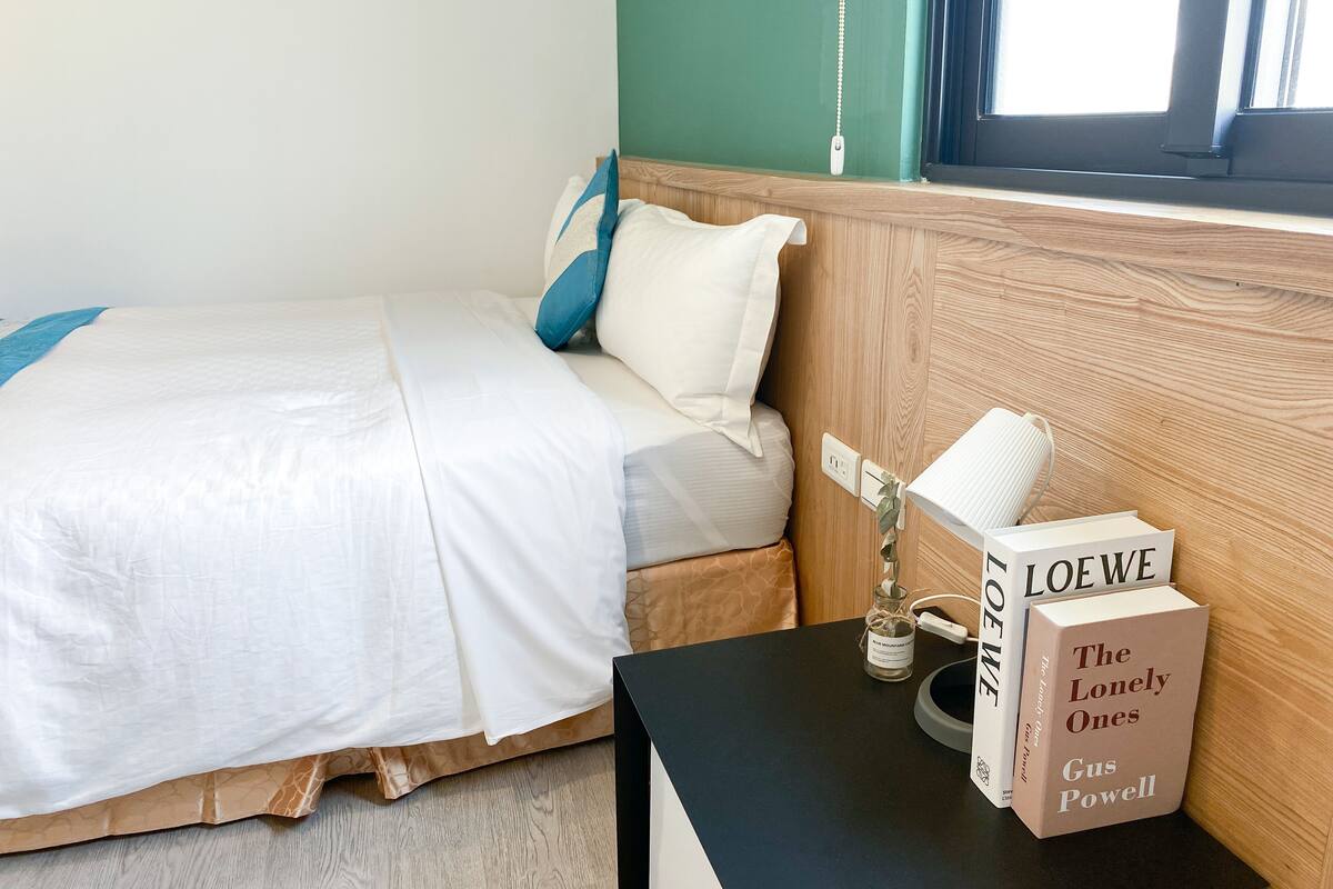 Un lit bien fait, avec une literie blanche et un oreiller bleu, une table de chevet sur laquelle repose un livre intitulé « Les Solitaires » de Gus Powell, et un petit vase avec une plante.