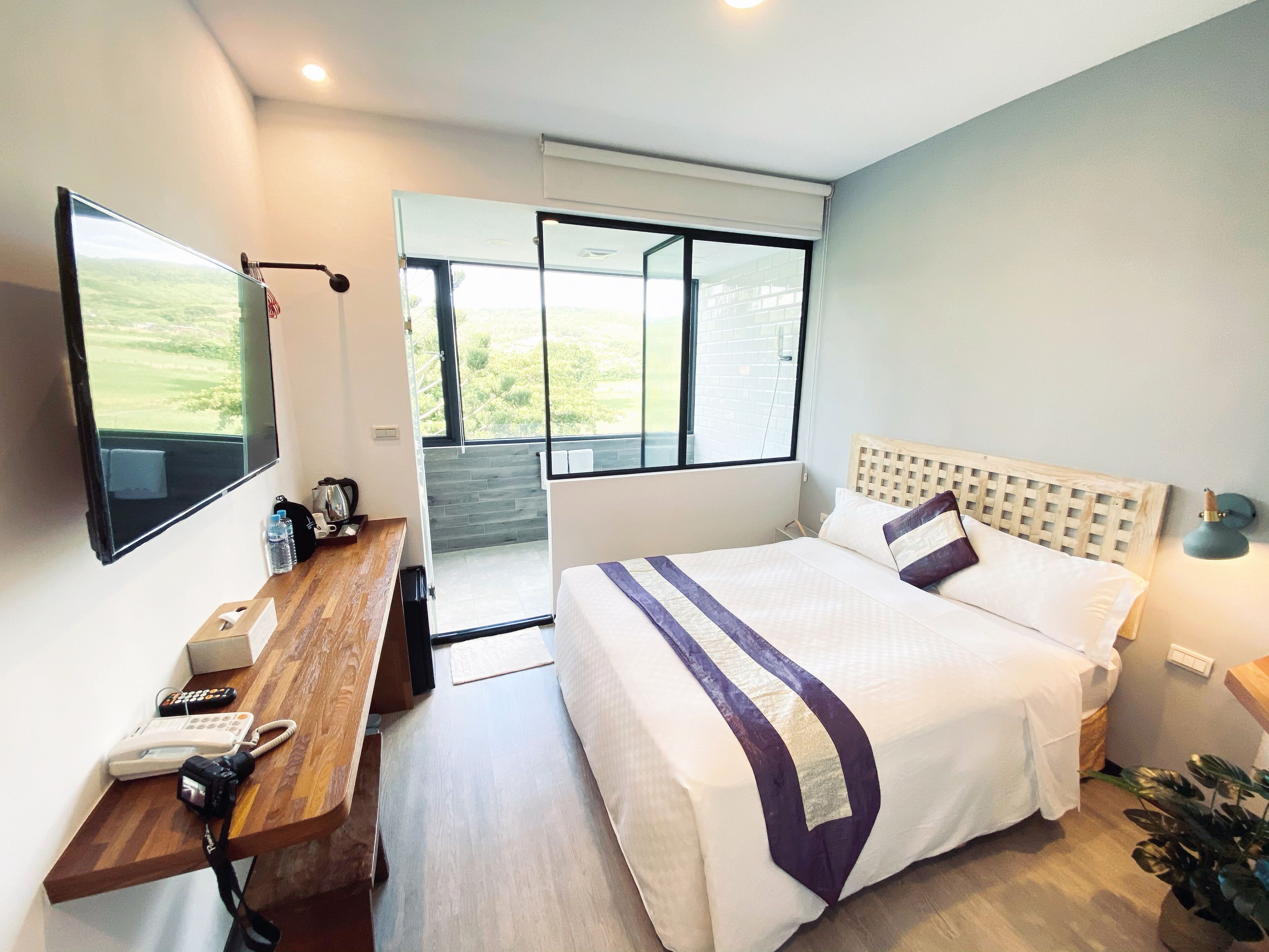 A modern hotel room with a large bed, a desk with a telephone and a camera, a window with a view of greenery, and a small plant.