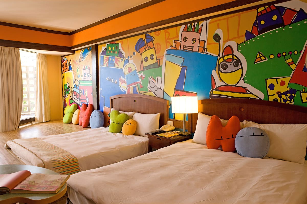 Habitación de hotel con dos camas, almohadas coloridas, una mesita de noche con lámpara y un mural en la pared.