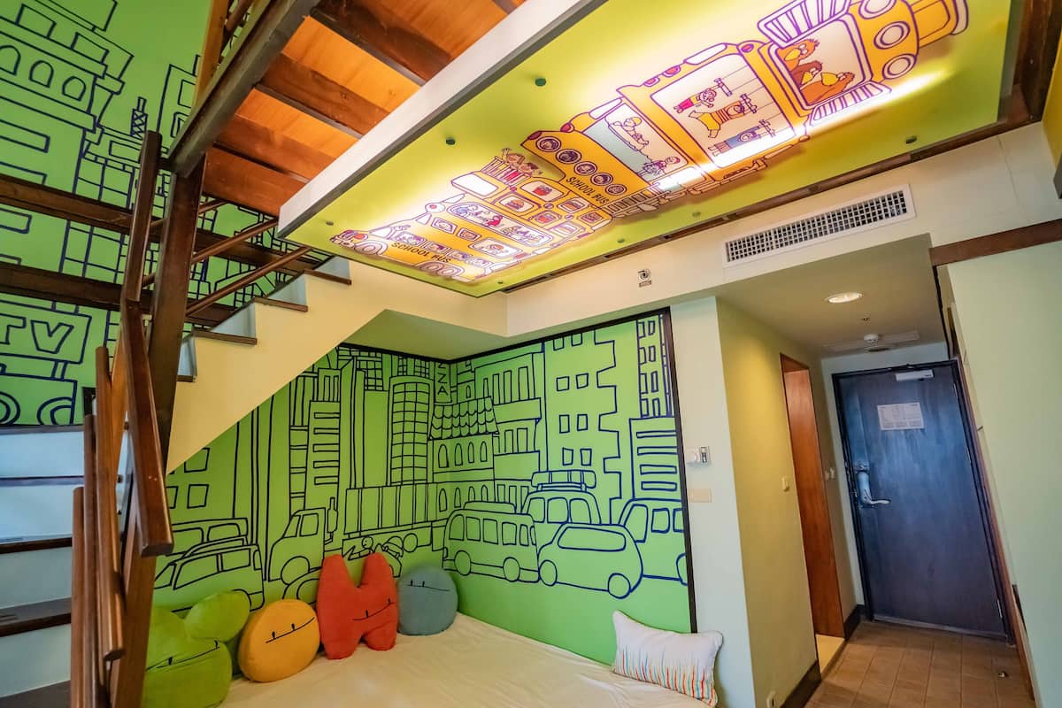 Una habitación con un mural de un paisaje urbano verde, una cama con almohadas y una escalera.