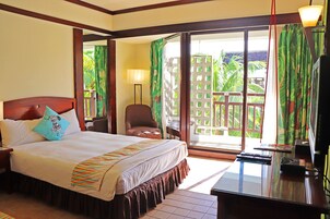 A hotel room with a large bed, a desk with a computer, a chair, and a balcony with a view of palm trees.