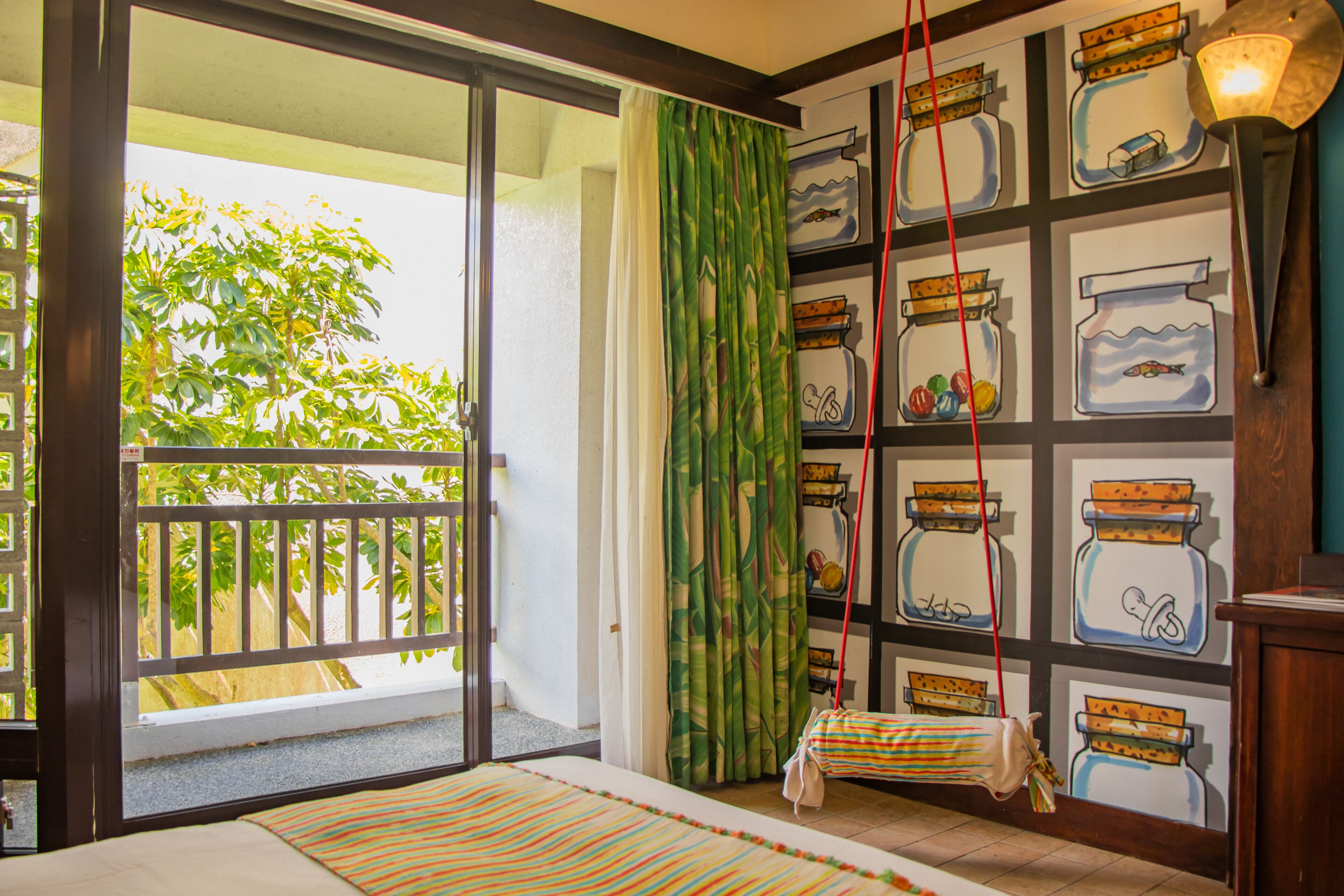 A room with a balcony, a sliding glass door, a bed with striped bedding, a hanging bag, and a wall with jars and a lamp.
