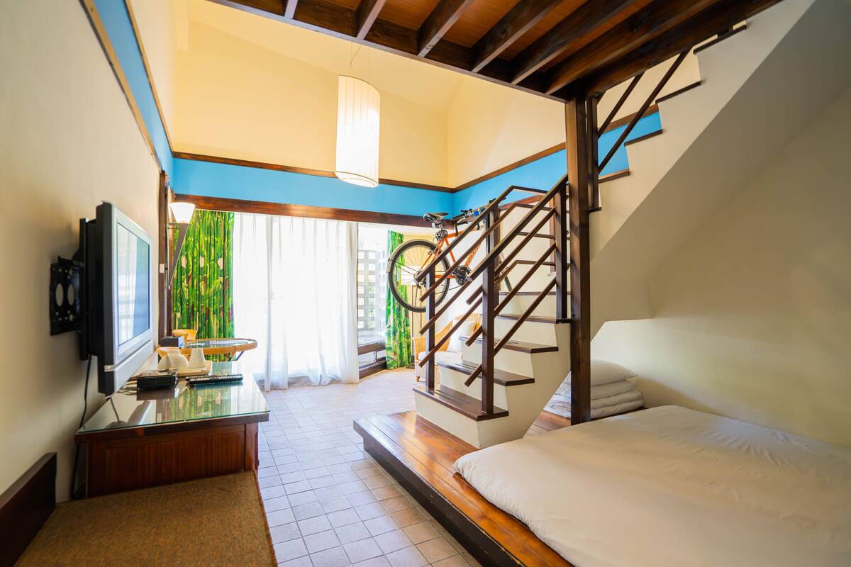 Habitación de hotel con cama, escritorio con televisor, escalera y vista a zonas verdes.