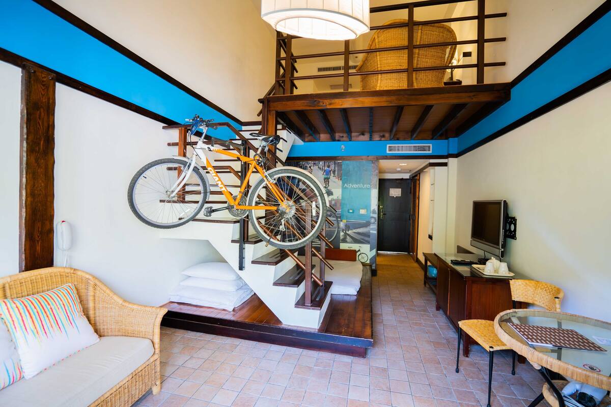 Una habitación con una escalera, una bicicleta colgada en la pared, un sofá, una mesa con sillas y un televisor.