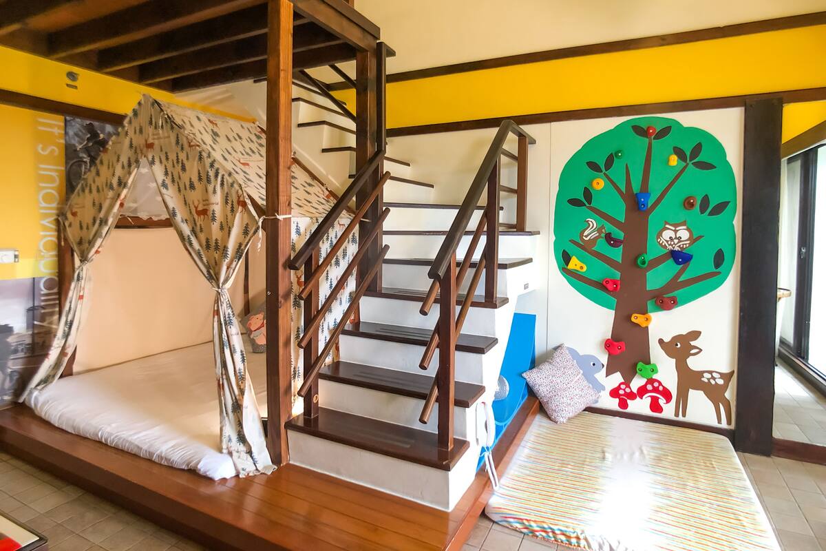 Una habitación con escalera de madera, una cama con manta a rayas y un mural en la pared con un árbol y animales.