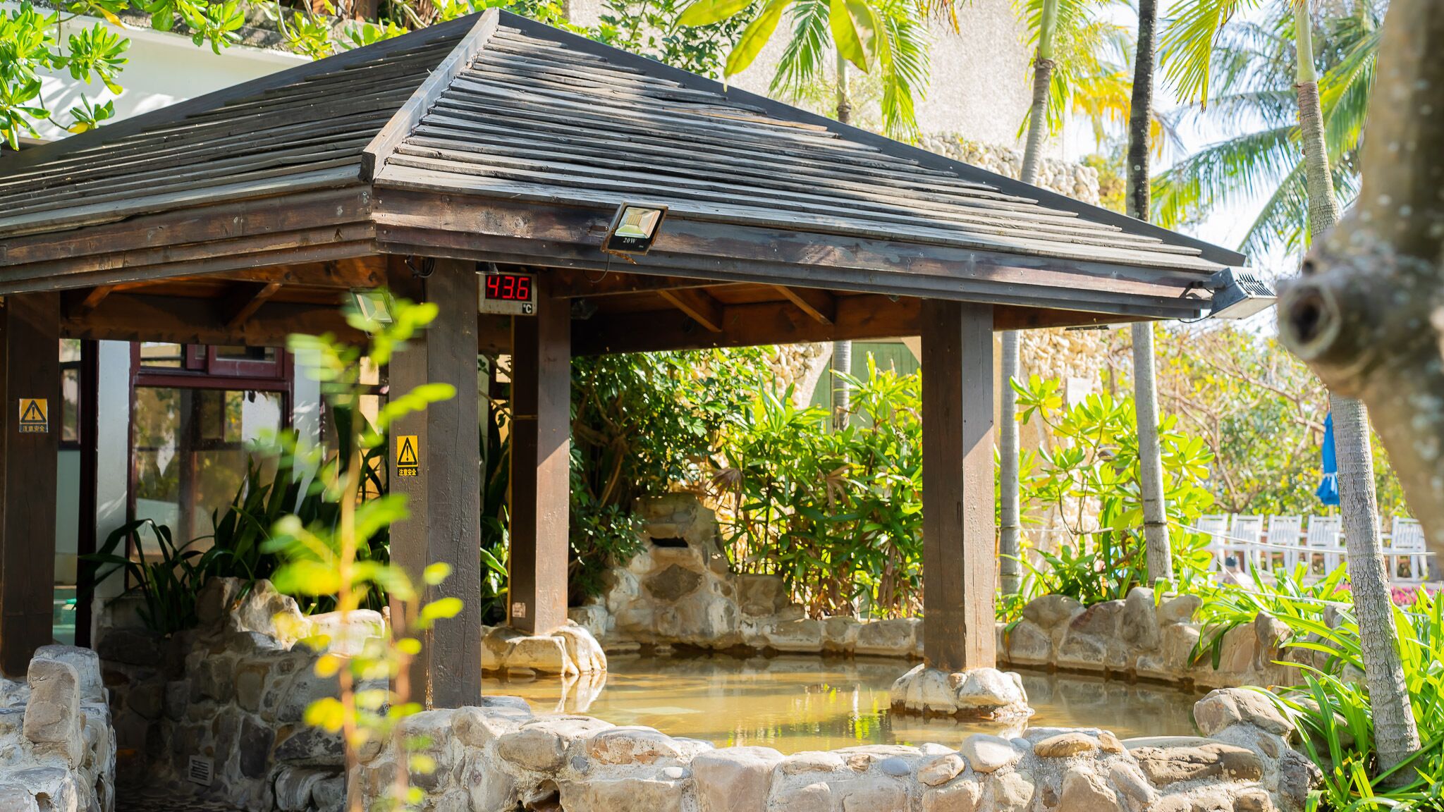 A wooden pavilion with a stone foundation, surrounded by lush greenery and a small pond.