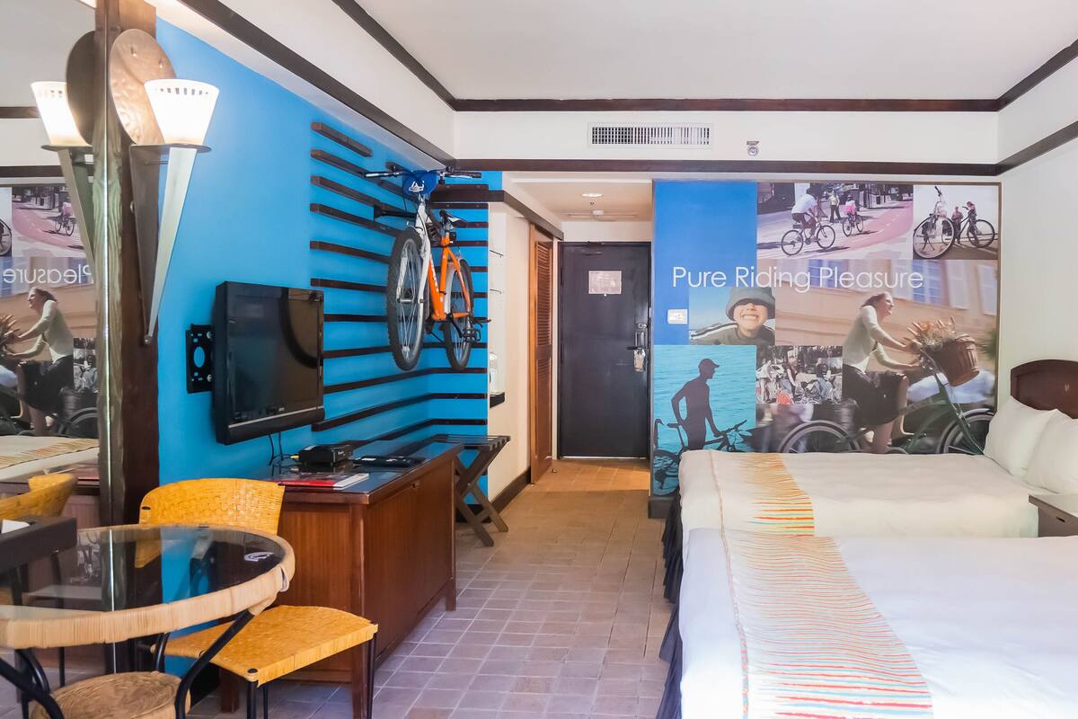 Habitación de hotel con cama, escritorio, silla, televisor y una pared decorada con una bicicleta y un póster.