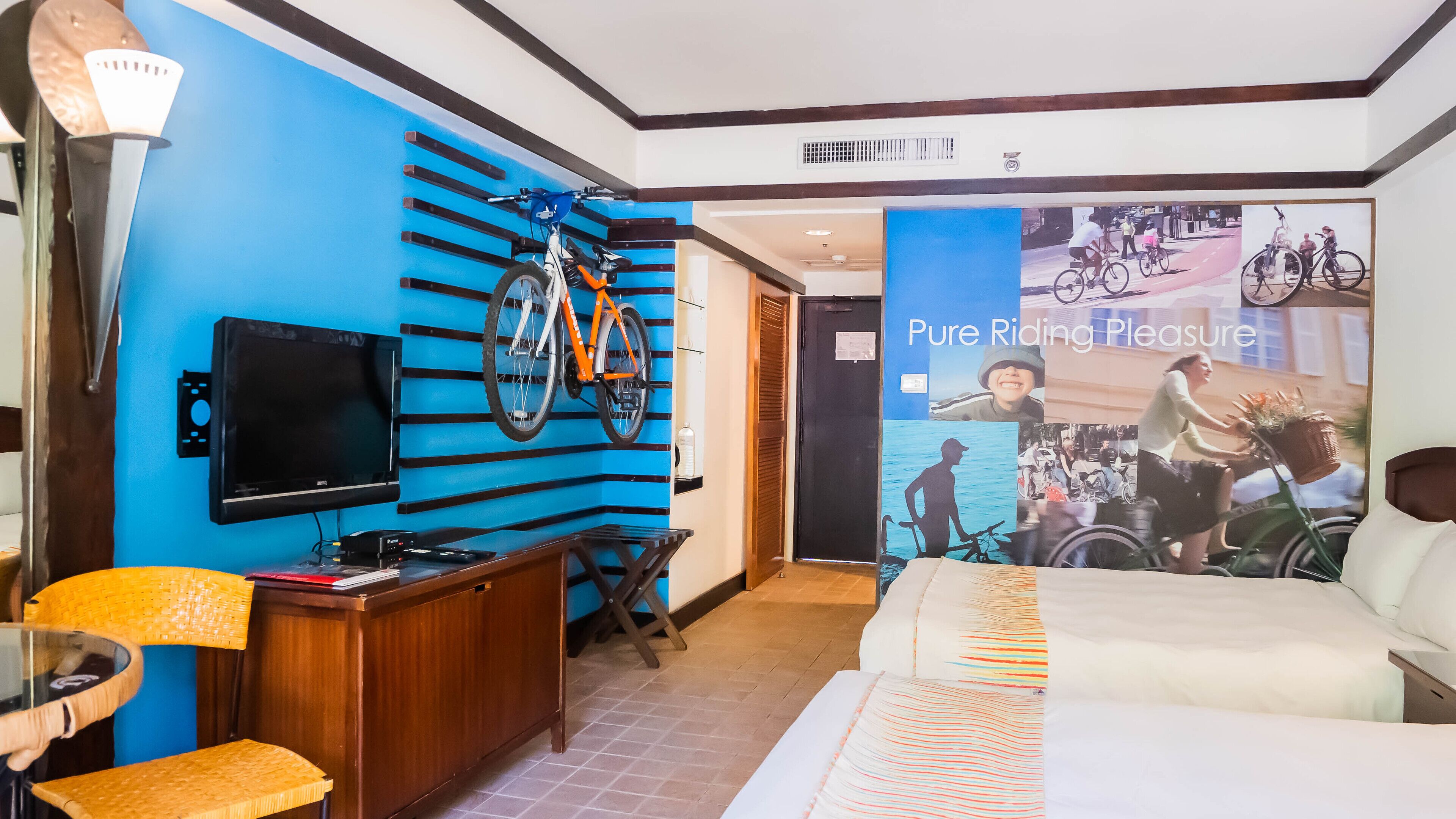 Habitación de hotel con pared azul, una bicicleta montada, televisor de pantalla plana, escritorio de madera, silla de mimbre y cama con colcha estampada.