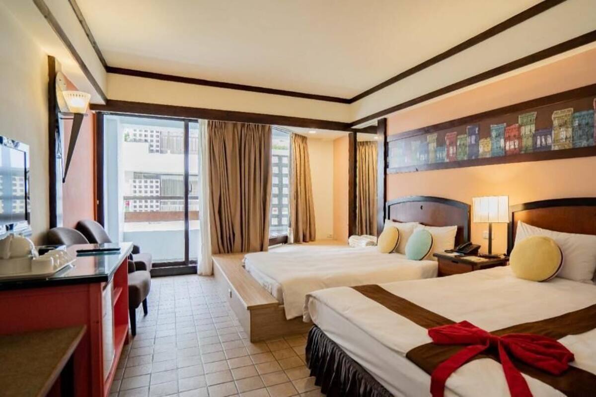Habitación de hotel con dos camas, un escritorio, una silla, una ventana con cortinas y una pared decorada con obras de arte coloridas.