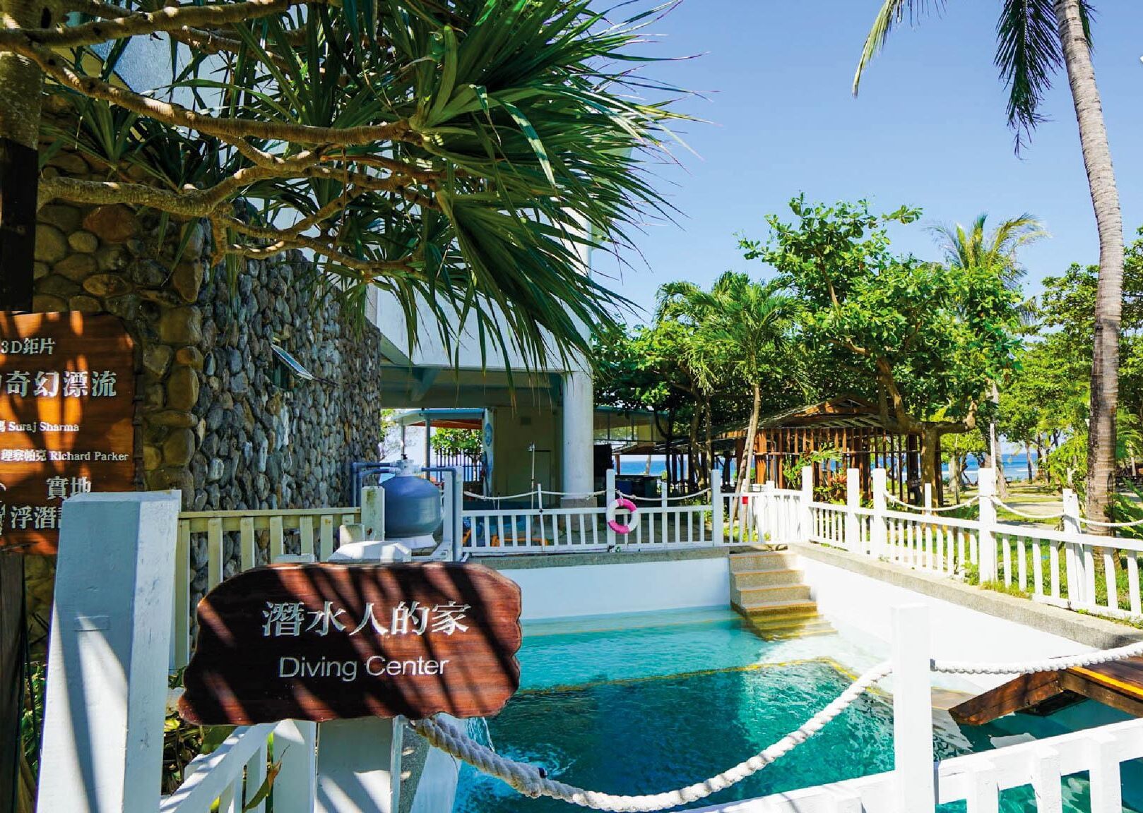 A diving center with a clear pool, surrounded by palm trees and a stone wall.