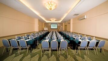 A conference room with rows of tables and chairs, a projector screen, and a ceiling light fixture.