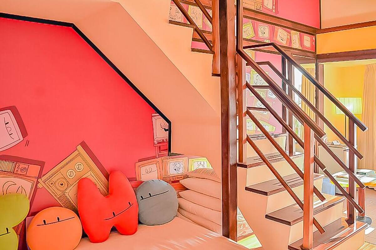 Un interior de colores vivos con una escalera, un sofá con cojines y una pared con un reloj y cuadros enmarcados.