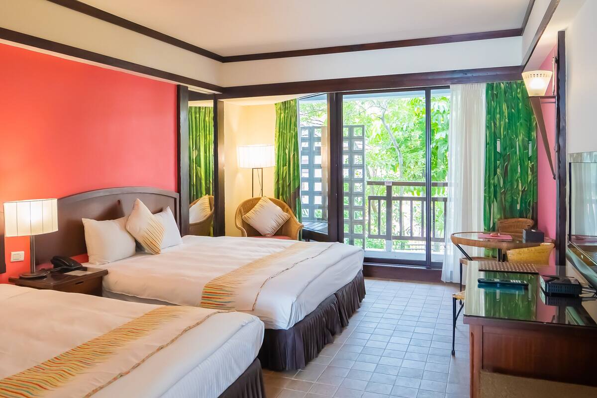 Habitación de hotel con dos camas, un escritorio con ordenador, una silla y vista a zonas verdes por la ventana.