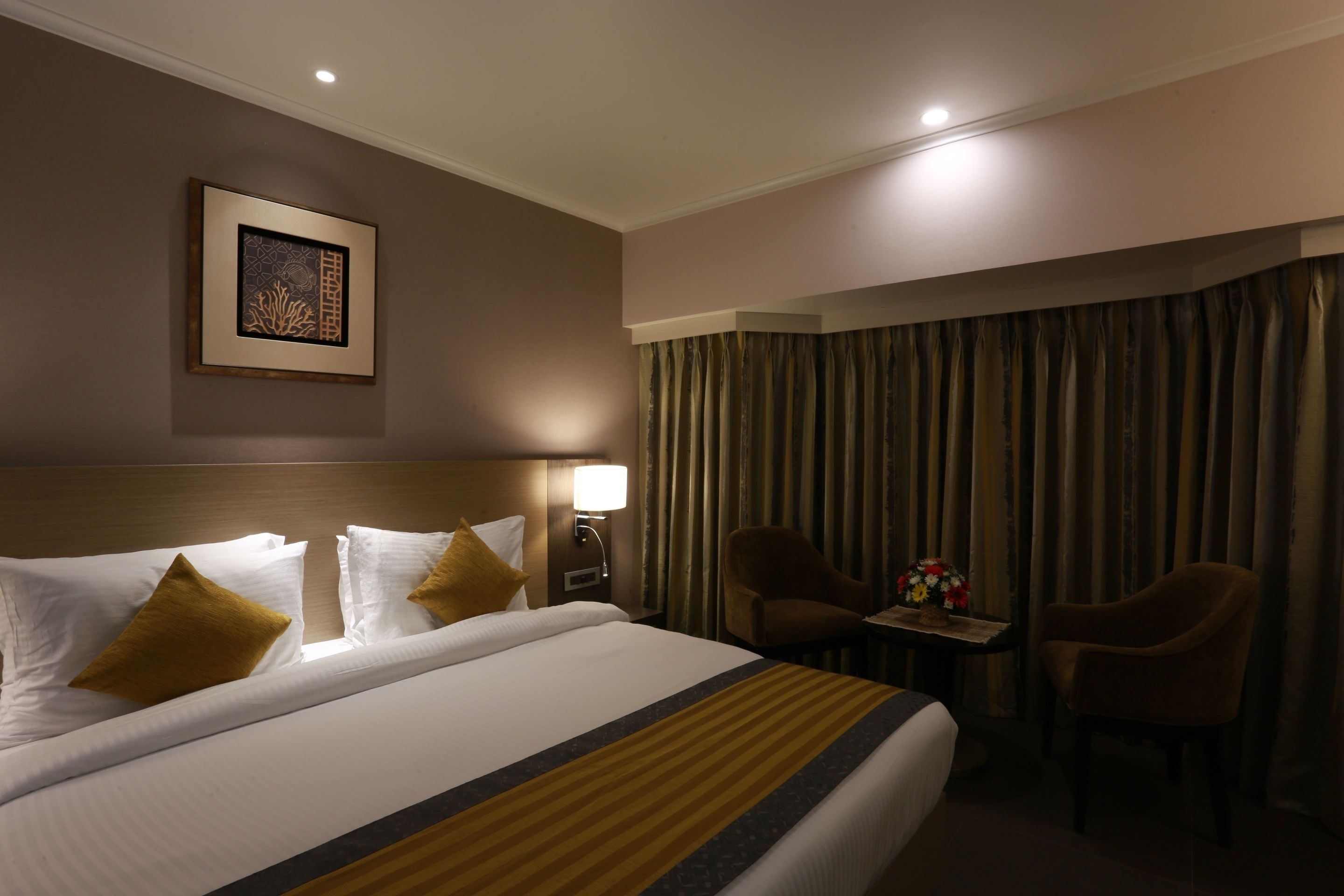 A hotel room with a large bed, two chairs, a small table with a vase, and a framed picture on the wall.