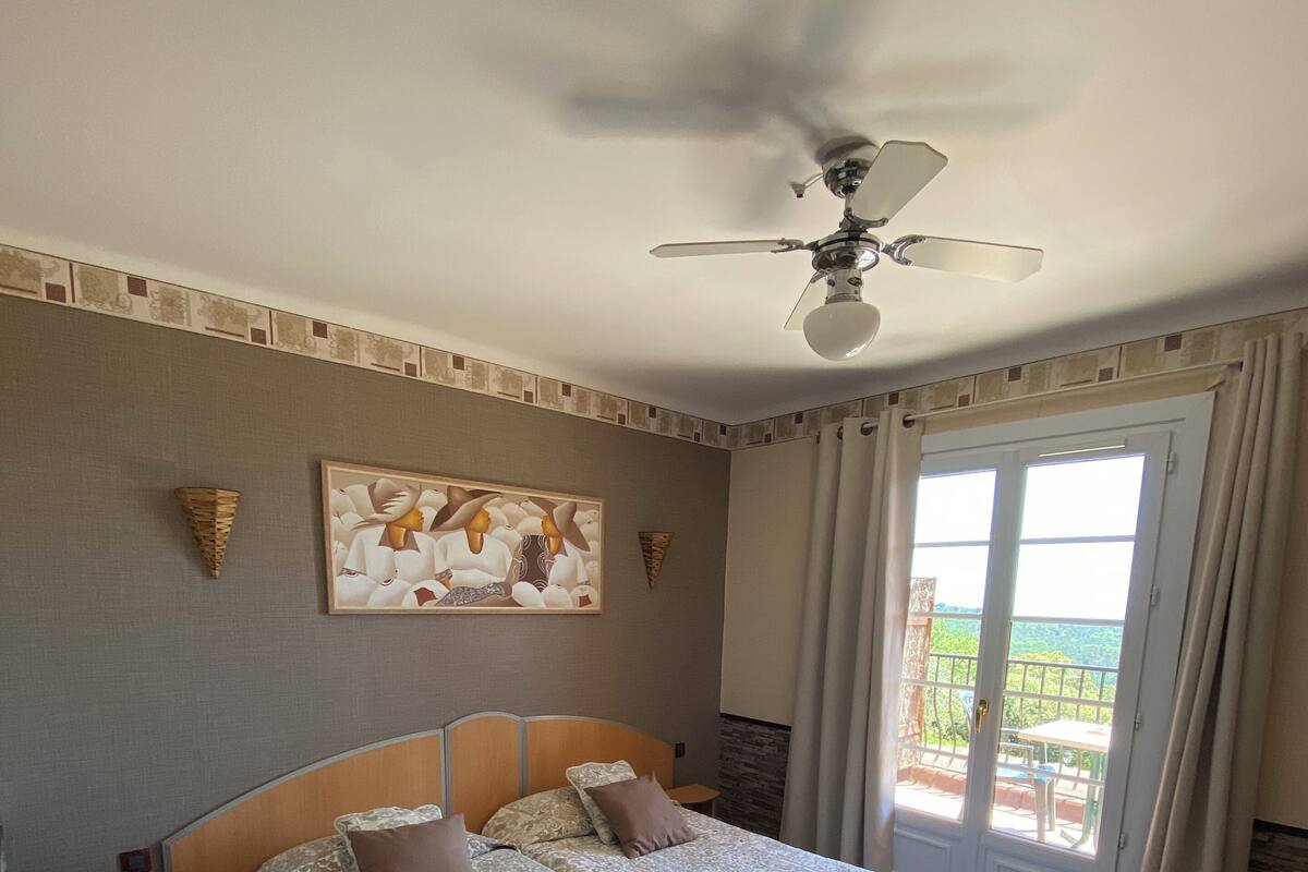 A hotel room with two beds, a ceiling fan, a painting on the wall, and a view of the outdoors through a sliding door.