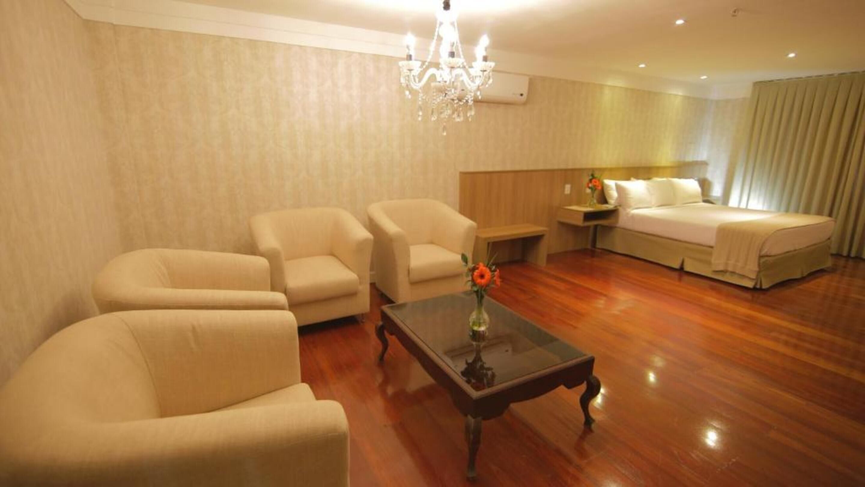 A hotel room with a wooden floor, a chandelier, a sofa set, a bed, and a nightstand with a vase of flowers.