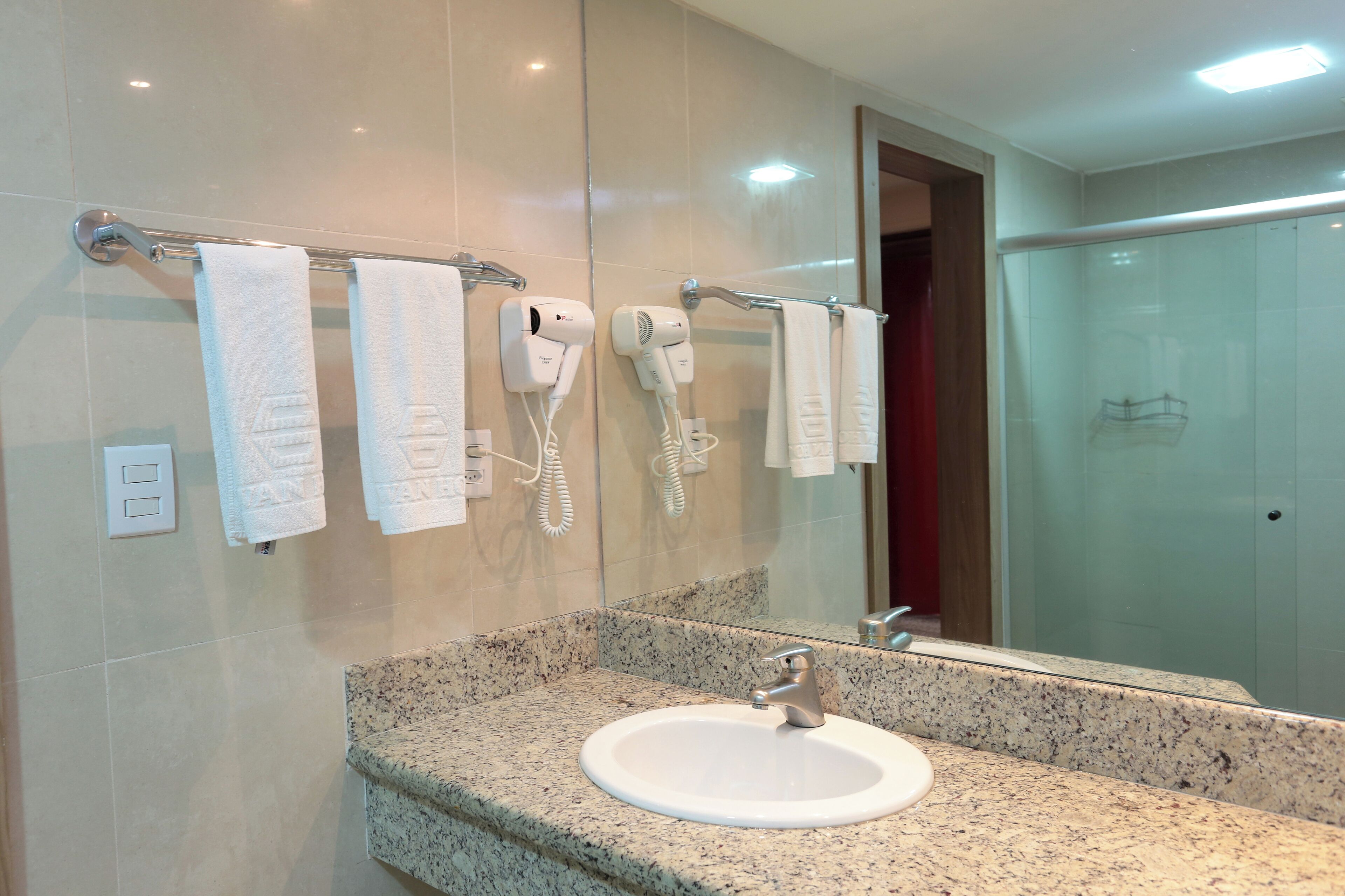 A bathroom with a granite countertop sink, two hair dryers mounted on the wall, and a glass shower enclosure.