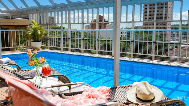 A poolside area with lounge chairs, a table with a drink, and a view of the cityscape. - Taiwan Hotel (Ribeirão Preto)