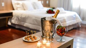 A neatly made bed with white linens, a bedside table with a champagne bottle, glasses, and a fruit bowl. - Taiwan Hotel (Ribeirão Preto)