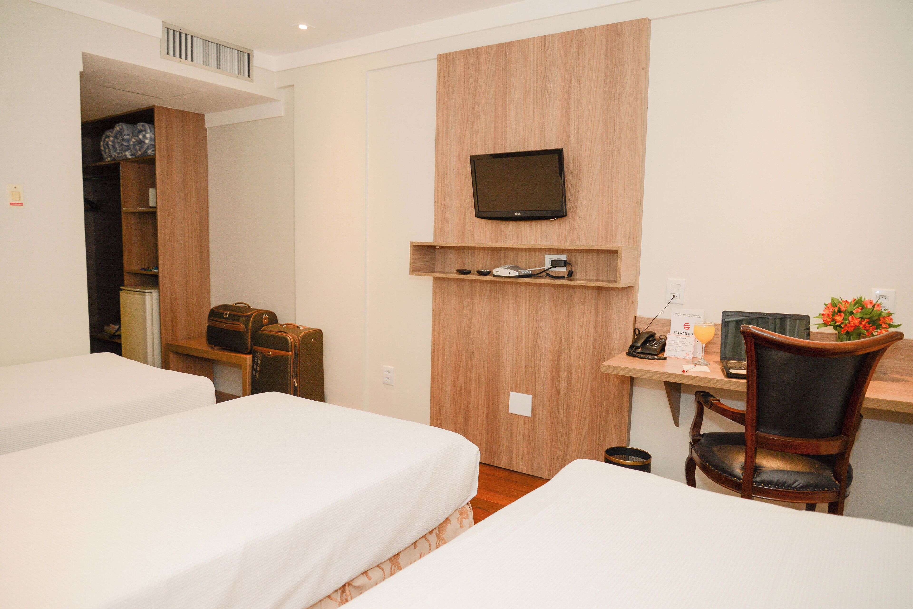 A hotel room with two beds, a desk with a computer and a chair, a TV mounted on the wall, and a small shelf with items.
