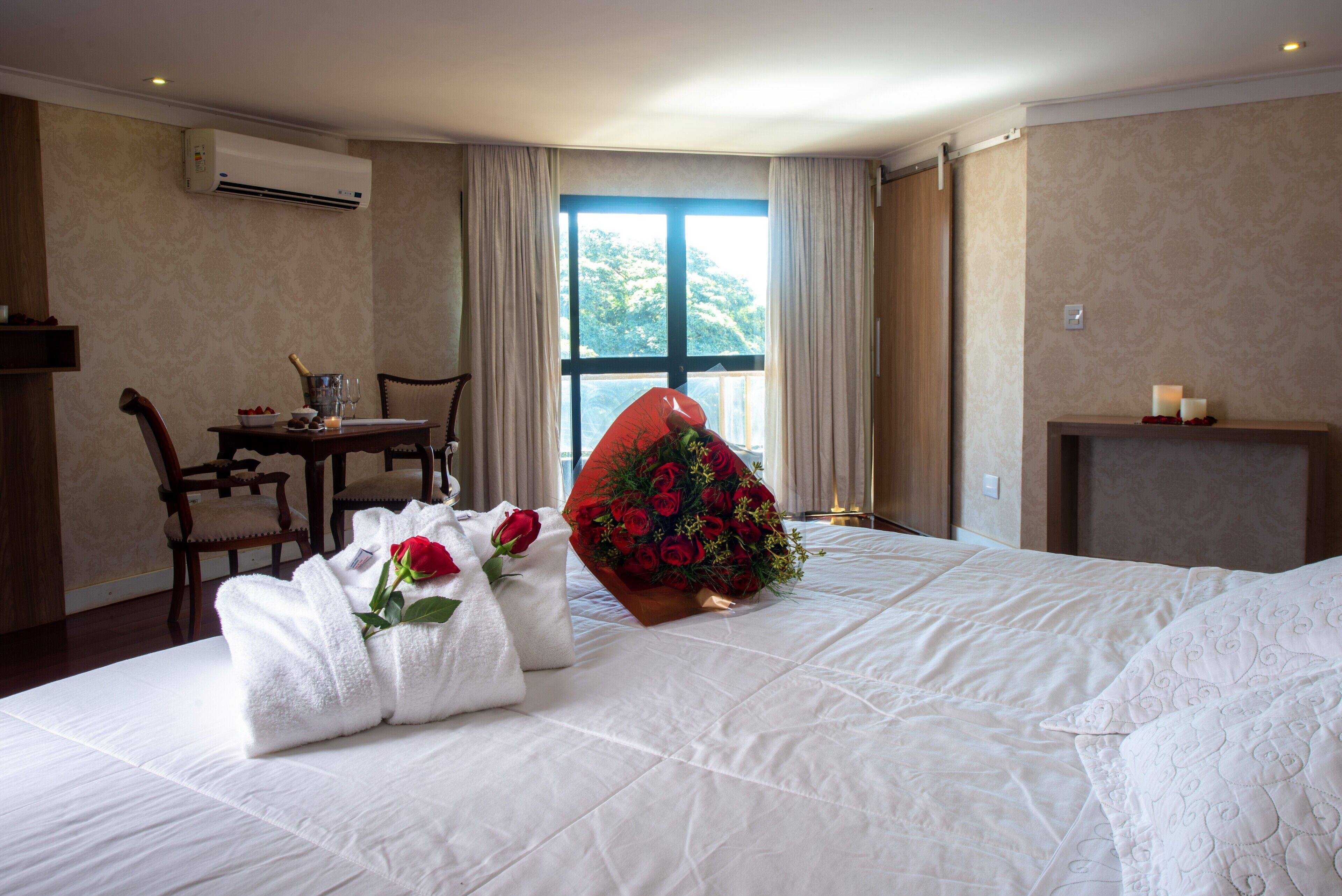 Een hotelkamer met een bed, bureau, stoel, raam met gordijnen en een boeket rode rozen.
