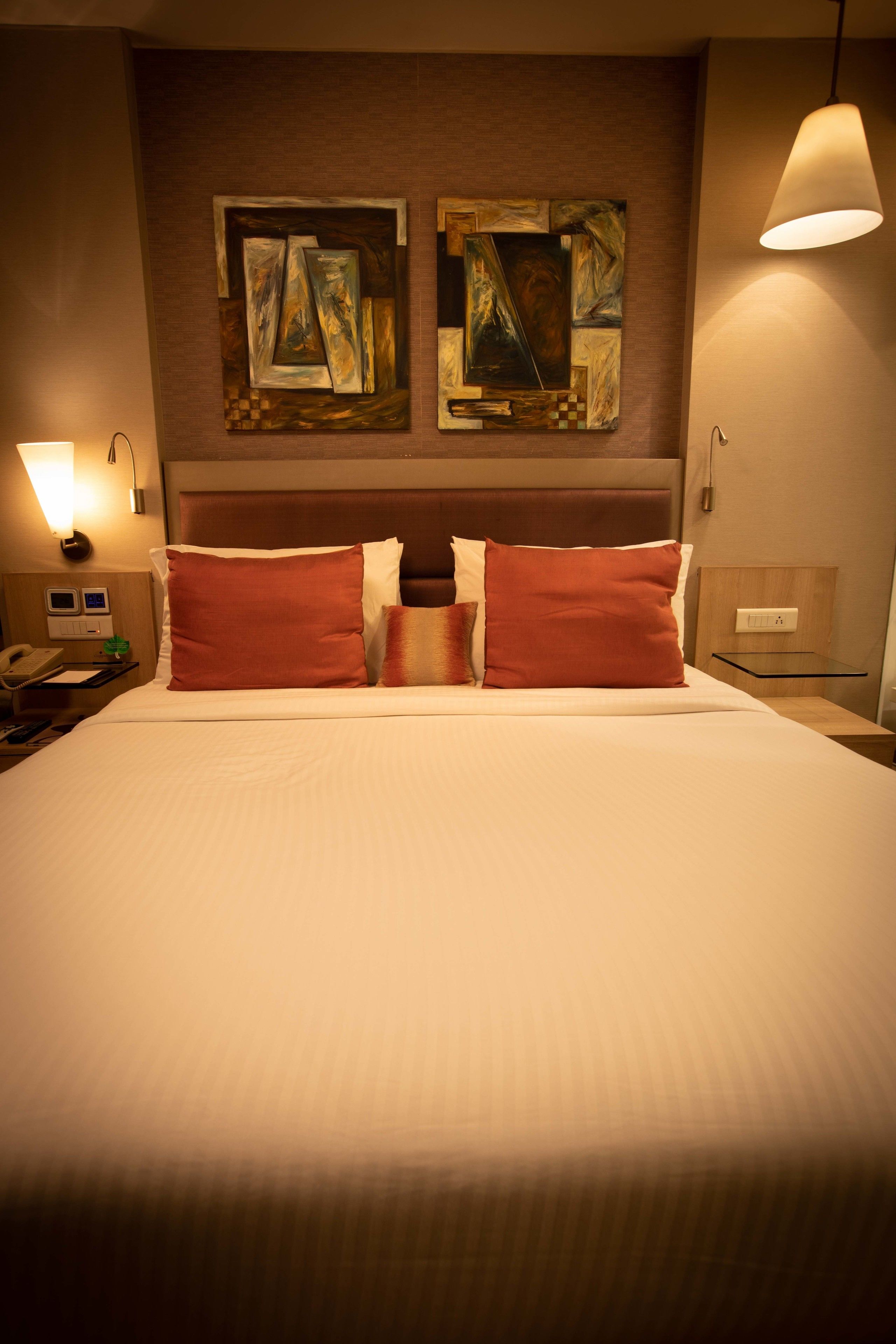 A hotel room with a large bed, two wall-mounted paintings, a bedside lamp, a nightstand, and a telephone.