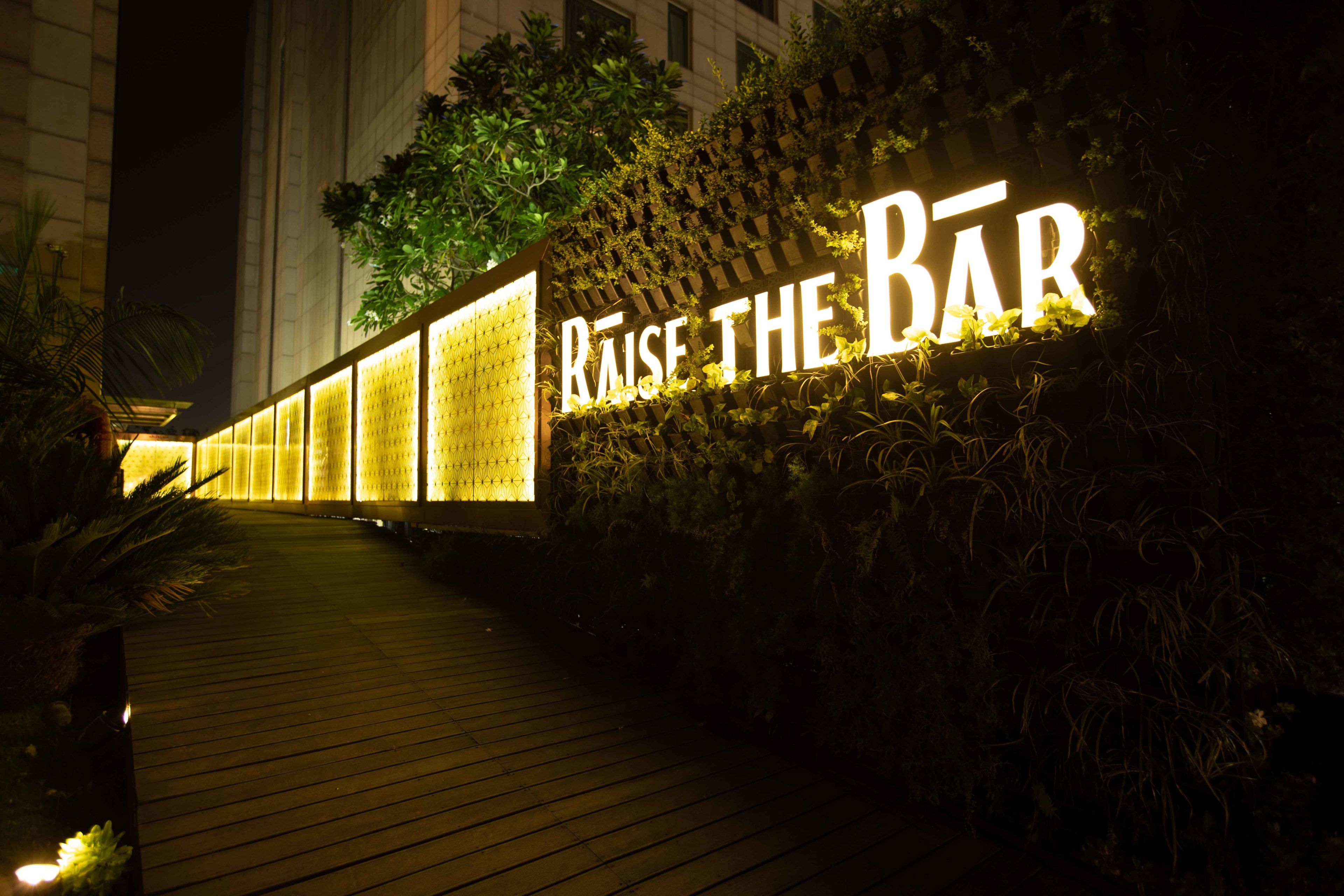 A nighttime view of a bar entrance with illuminated signage reading "RAISE THE BAR" and a wooden pathway leading to it.