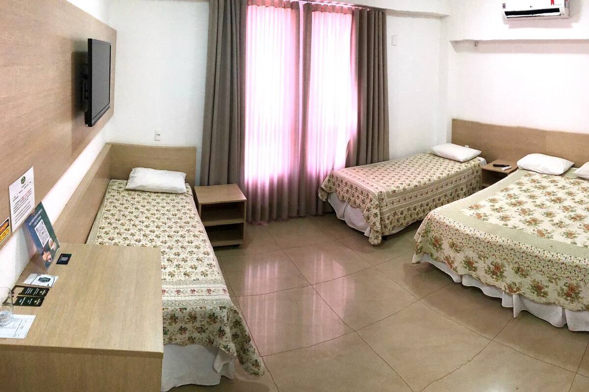 Habitación de hotel con dos camas individuales, un escritorio de madera, un televisor de pantalla plana, una ventana con cortinas y suelo de baldosas.