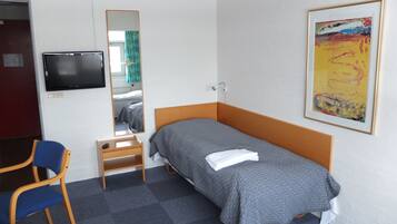 A single-bed room with a wooden headboard, a small bedside table with a remote control, a blue chair, a mirror, a TV mounted on the wall, and a framed abstract painting.