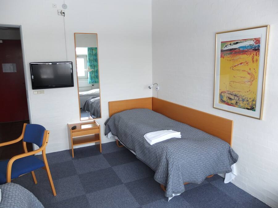 A single-bed room with a wooden headboard, a small bedside table with a remote control, a blue chair, a mirror, a TV mounted on the wall, and a framed abstract painting.