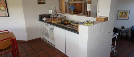 Una cocina con mostrador blanco, un lavamanos y piso de baldosas. Se ven sillas y una mesa en primer plano.