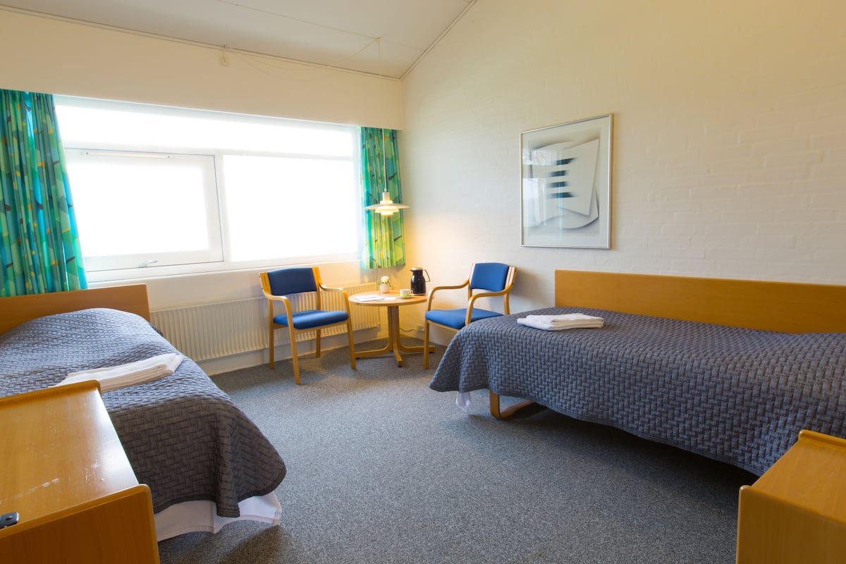 A hotel room with two single beds, a small round table with a chair, a window with curtains, and a framed picture on the wall.