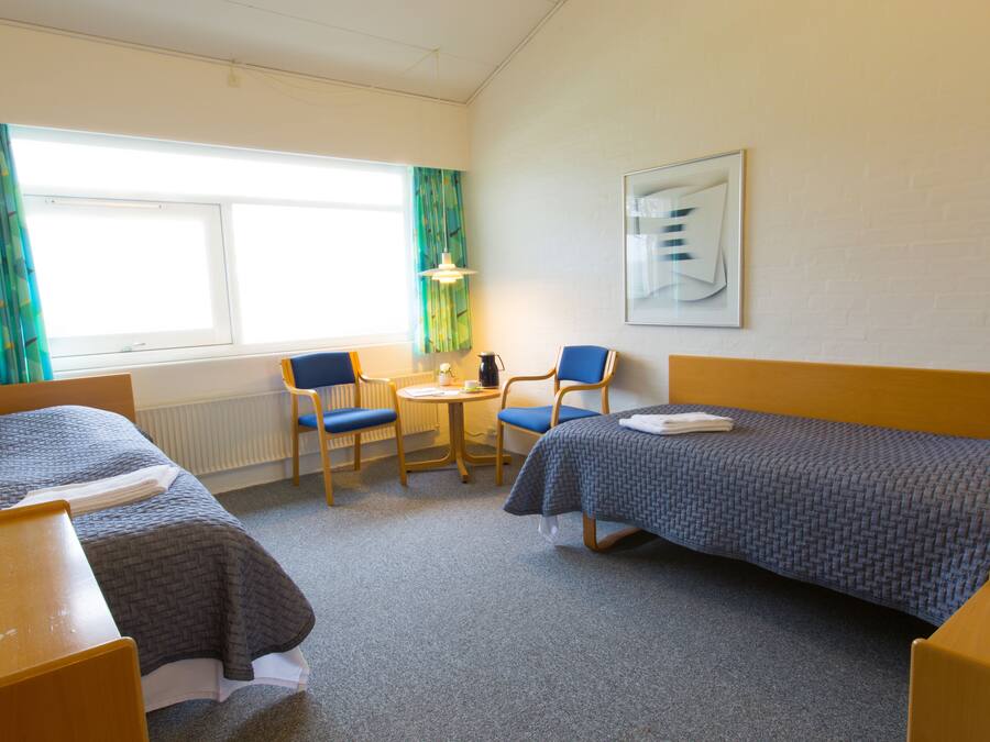 A hotel room with two single beds, a small round table with a chair, a window with curtains, and a framed picture on the wall.