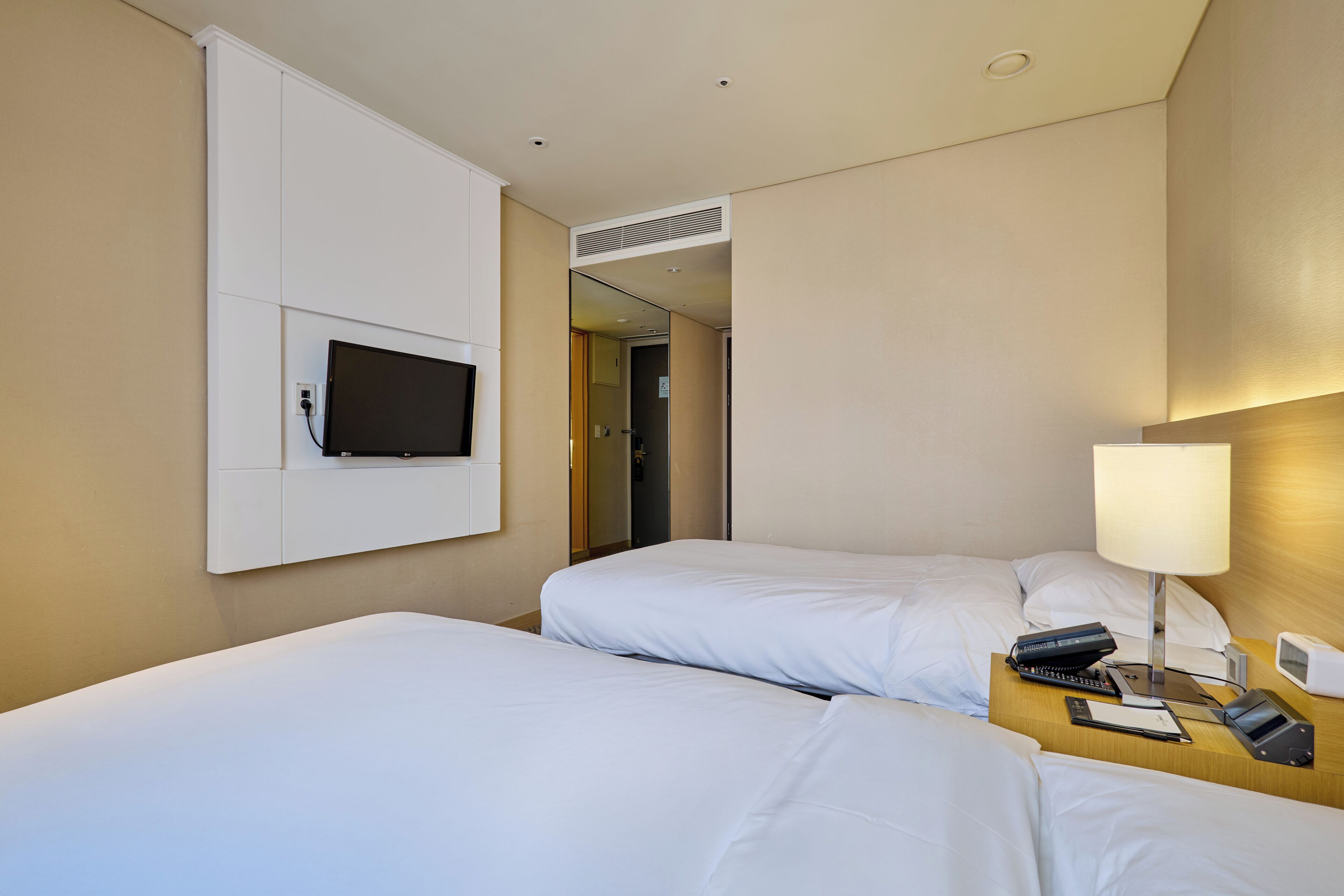 A hotel room with two beds, a TV mounted on the wall, a bedside table with a lamp, and a clock.