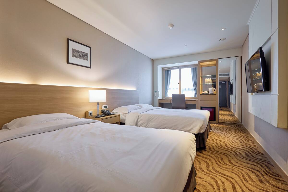 Une chambre d’hôtel avec deux lits, un bureau, une chaise, une télévision et une salle de bain visible à travers une porte ouverte.
