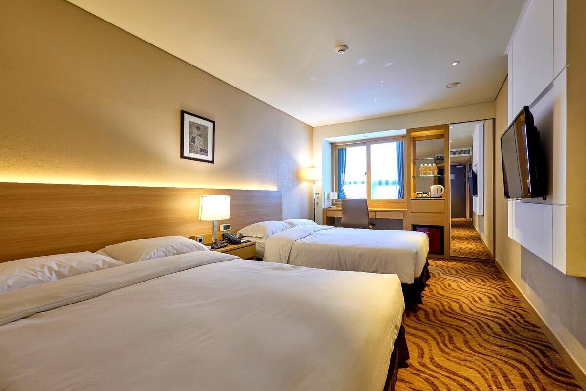 Une chambre d’hôtel avec deux lits, un bureau avec une chaise, une télévision et une salle de bain visible à travers une porte en verre.