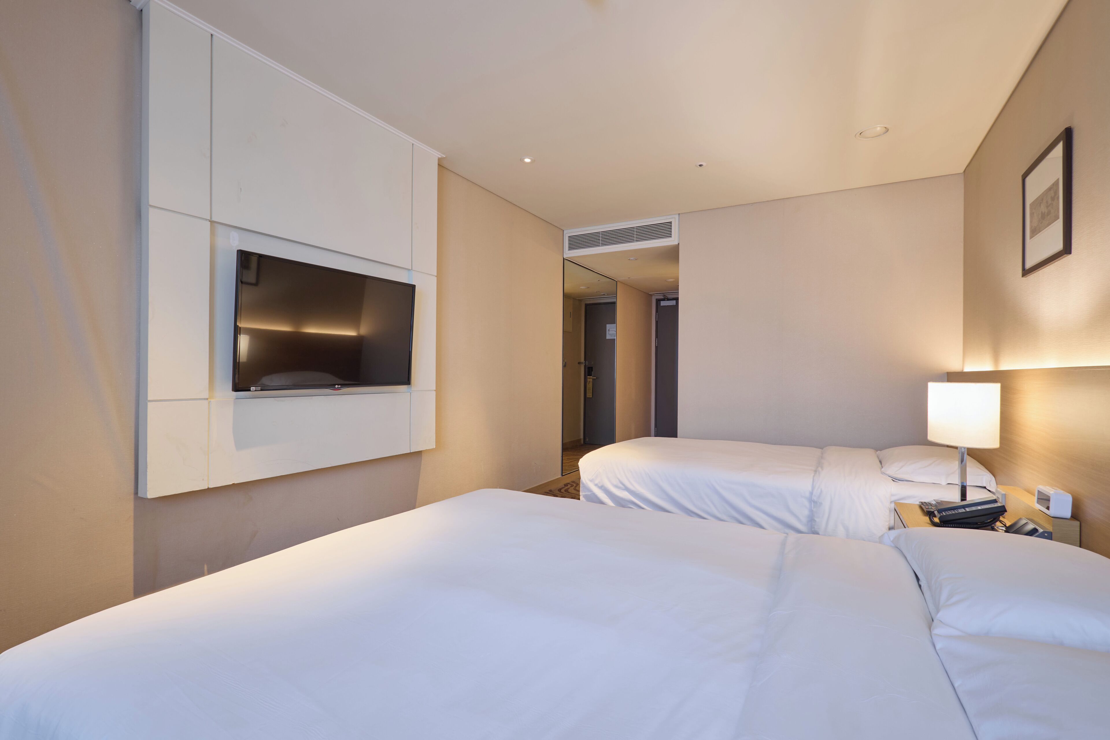 Habitación de hotel con dos camas, un televisor montado en la pared, una lámpara de noche y una puerta que da a otra habitación.