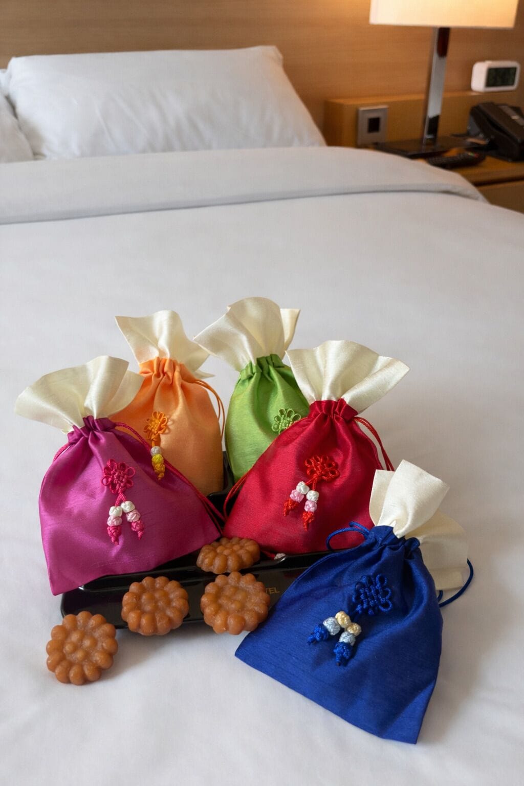 A bed with white linens, a bedside table with a lamp, and a tray with colorful drawstring bags and decorative items.