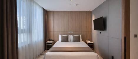 A hotel room with a large bed, a TV mounted on the wall, and a window with curtains.