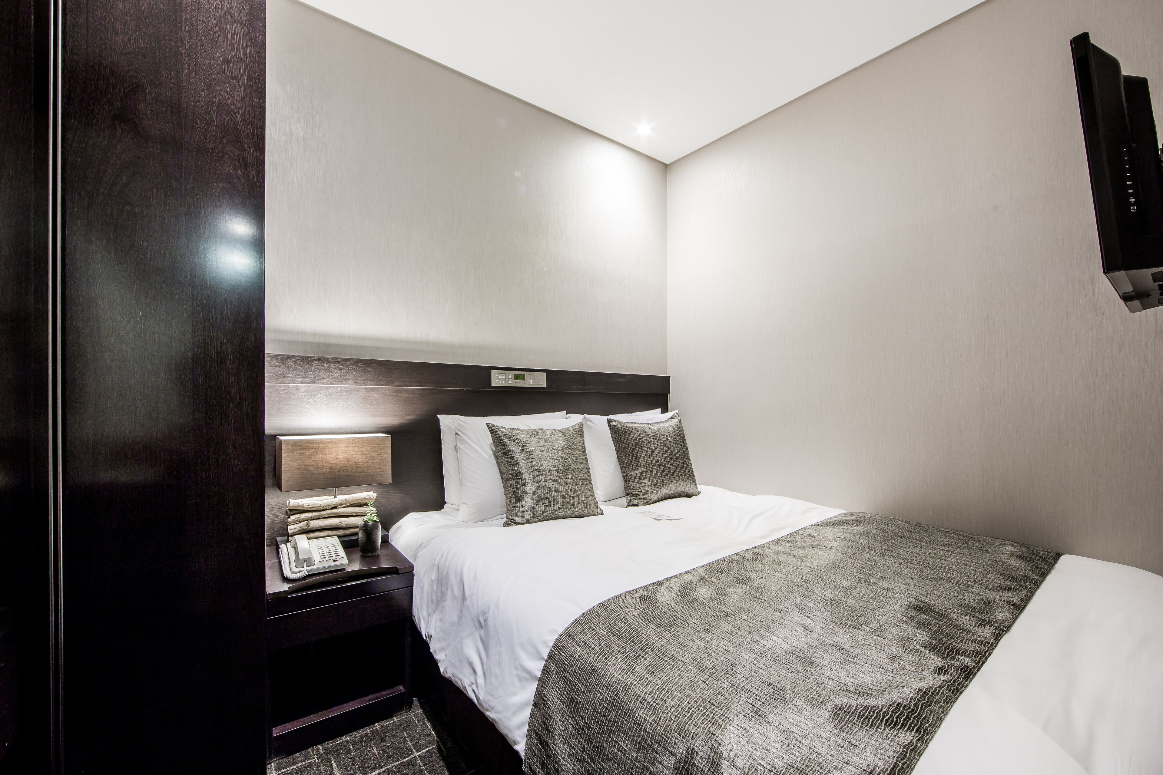 A modern hotel room with a neatly made bed, a bedside table with a lamp, and a wall-mounted TV.