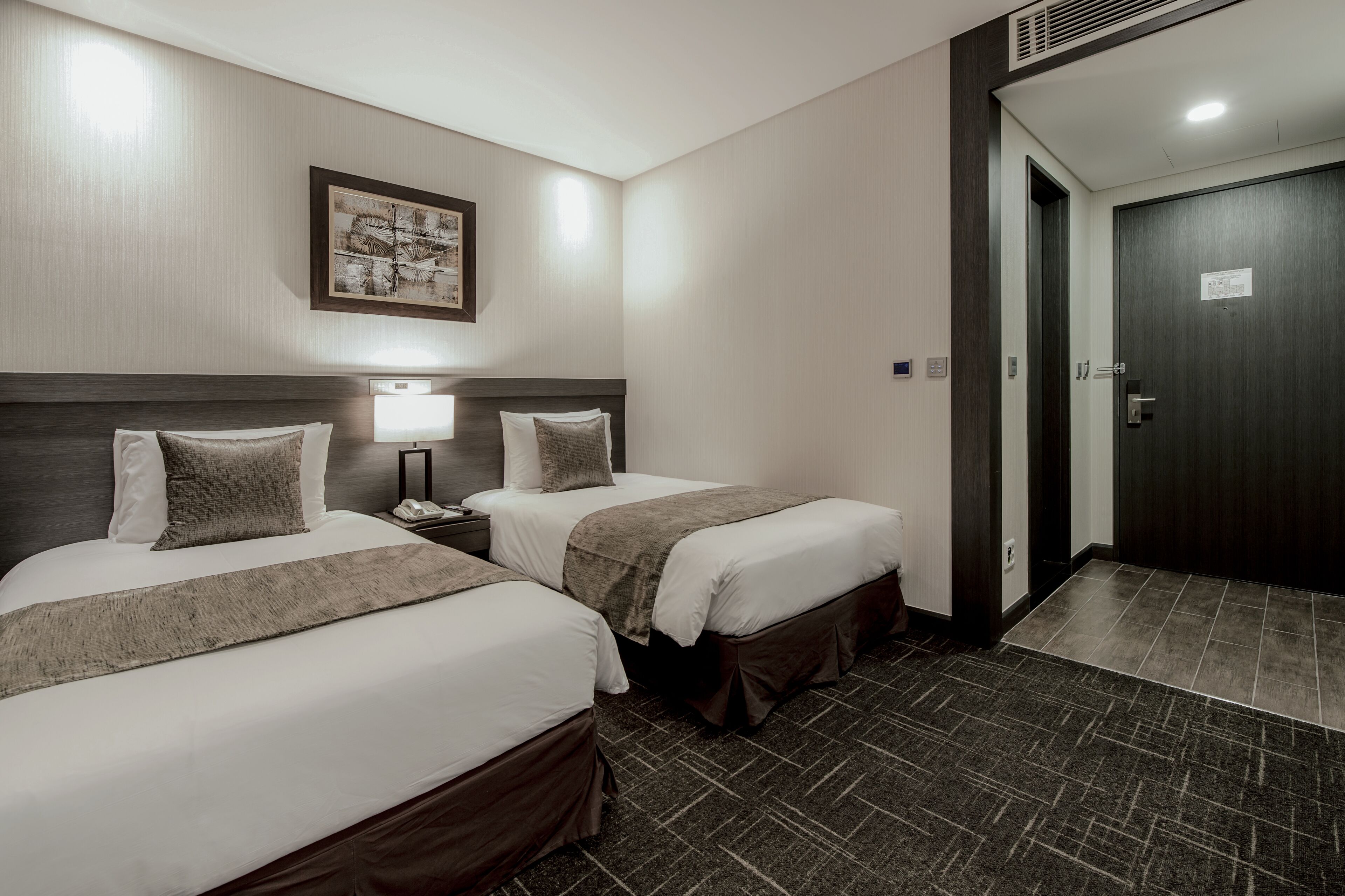 A hotel room with two beds, a nightstand, a lamp, a framed picture on the wall, and a door with a sign.
