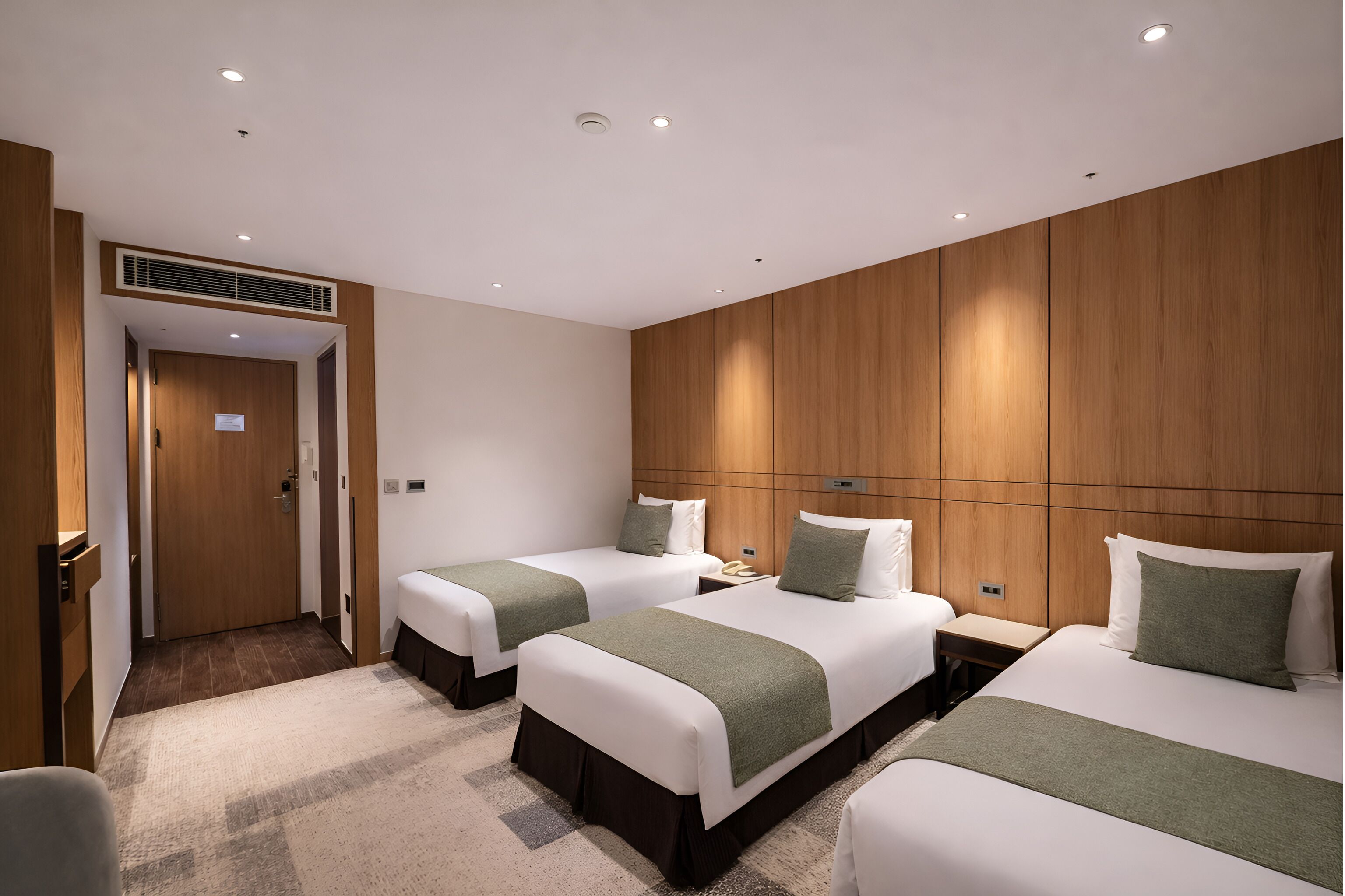 Een hotelkamer met drie bedden, houten lambrisering aan de wanden, een vloerbedekking en een deur die naar een andere kamer leidt.