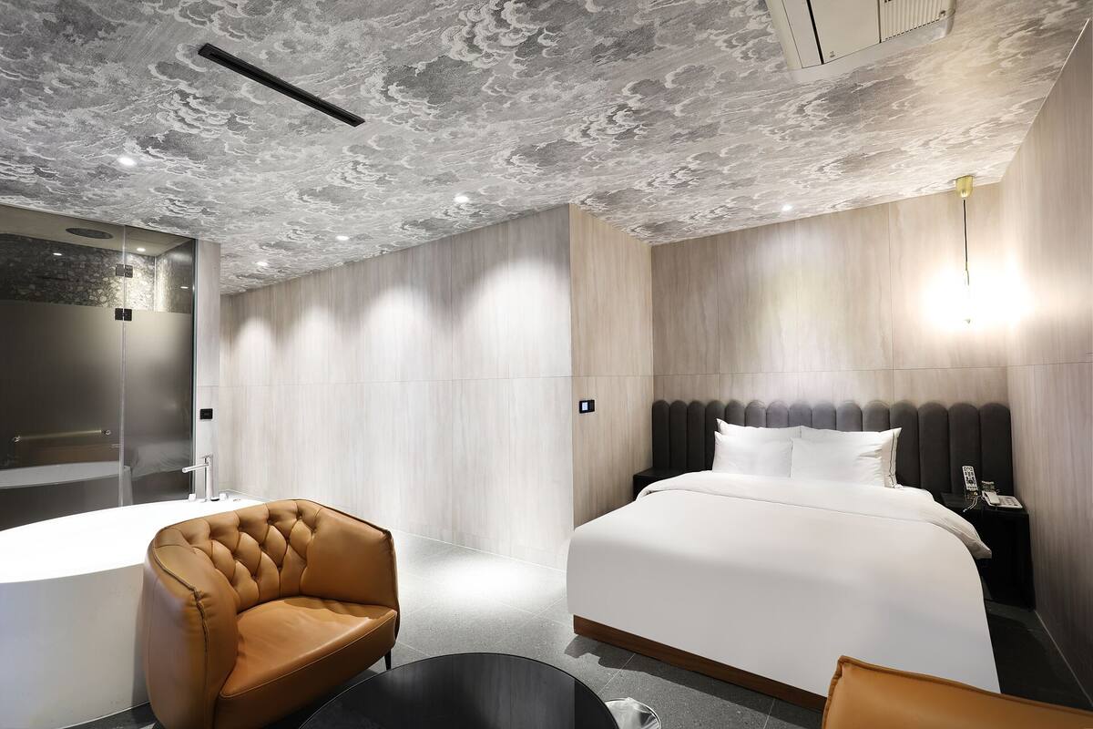Una habitación de hotel moderna con una cama grande, un sillón de cuero, una mesa redonda negra y una zona de ducha.