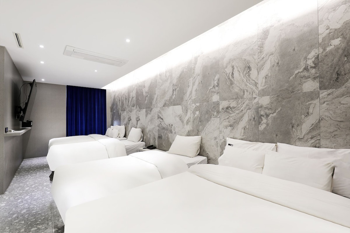 Habitación de hotel con pared de mármol, televisor de pantalla plana, cortina azul y varias camas.