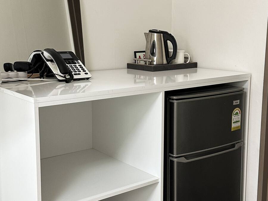 A compact hotel room with a white desk, a black refrigerator, a coffee maker, a phone, and a pair of black slippers.