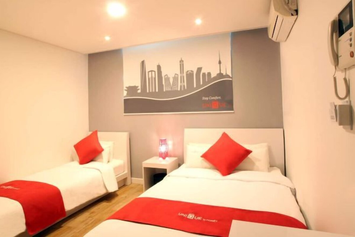 A hotel room with two beds, each with white bedding and red accents, a city skyline mural on the wall, and a bedside table with a lamp.